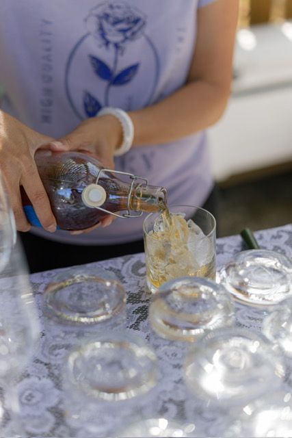 Person pouring amber liquid from a bottle into a glass of ice. Several empty glasses are on a lace-covered table.