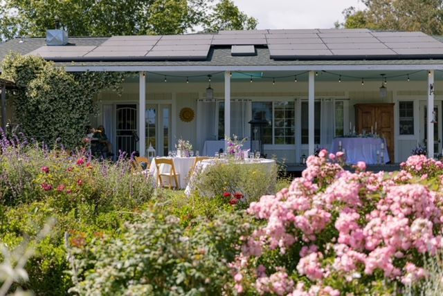 House with solar panels, a garden, and tables set for a gathering, with pink flowers in the foreground.