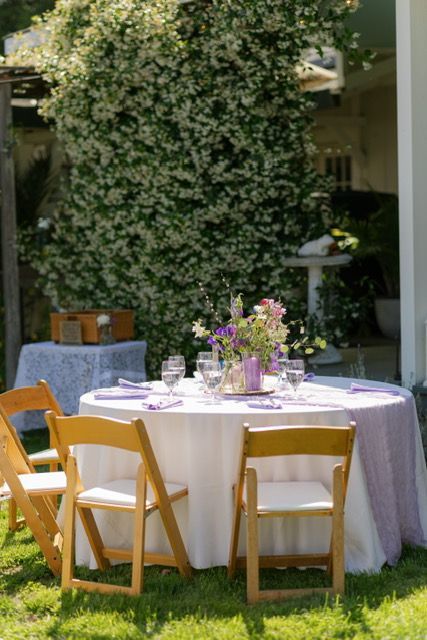 Round table set for a meal outdoors with chairs. Flowers and purple accents on white tablecloth.