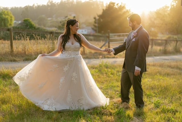 Couple holding hands outdoors, the bride in a flowing gown. Golden sunlight bathes the grassy field.