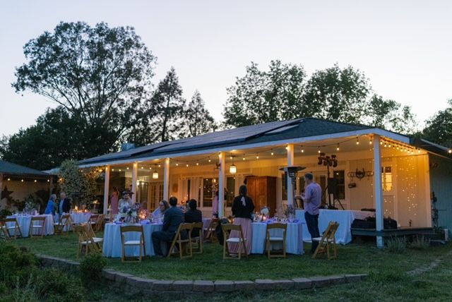 Outdoor evening event with tables, guests, and a building lit by string lights.