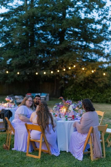 Guests at a table outdoors, lit by string lights, with purple flowers and tablecloth.