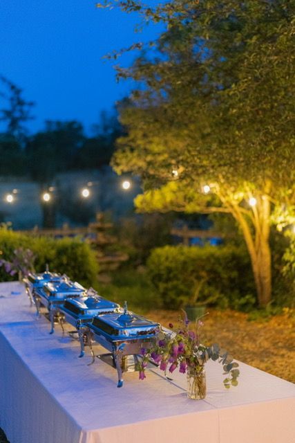 Buffet table outdoors at dusk with lit string lights and floral arrangement.