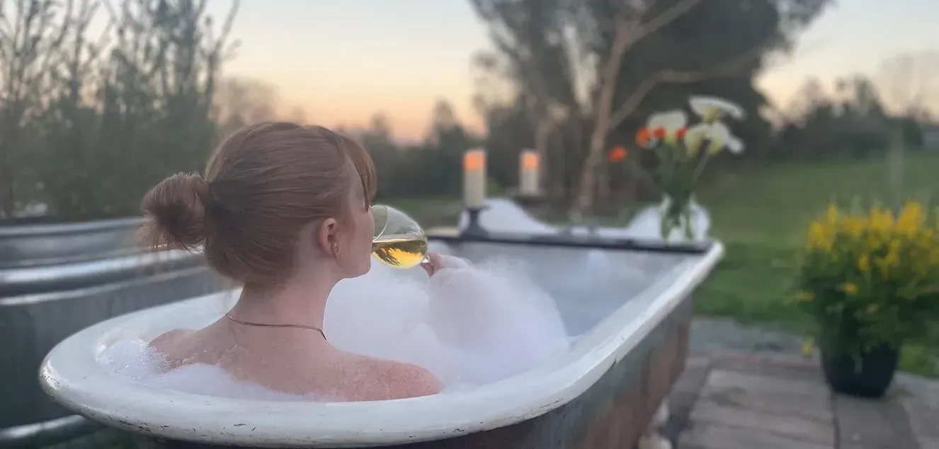 Person enjoys a bath outdoors, holding a glass of white wine. Soft focus with bubbles and candles.