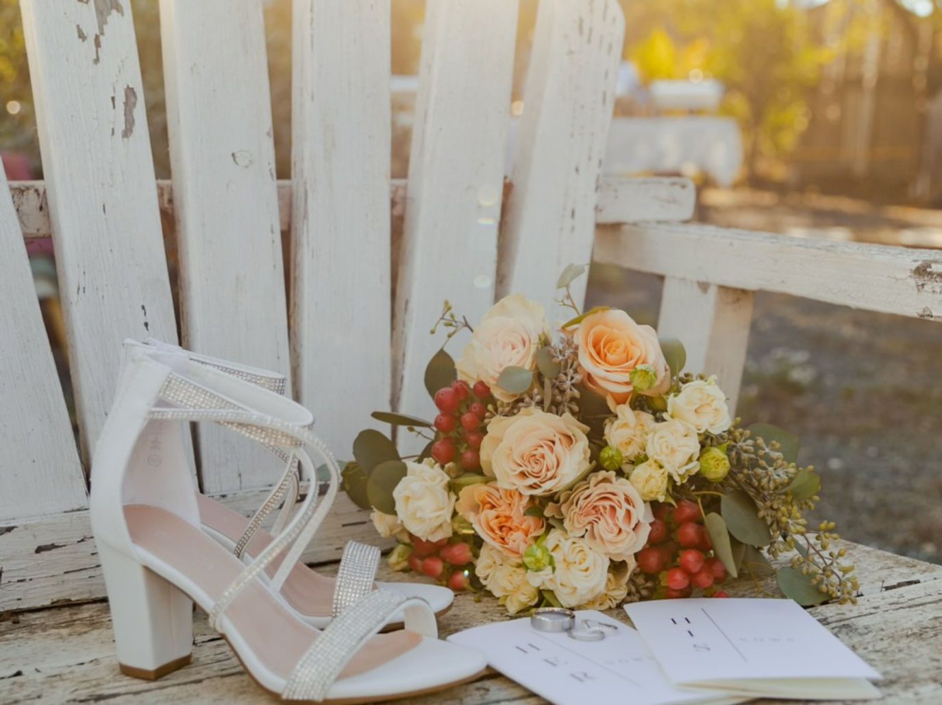 White wedding shoes, floral bouquet, rings, and invitation on a rustic, white wooden bench.