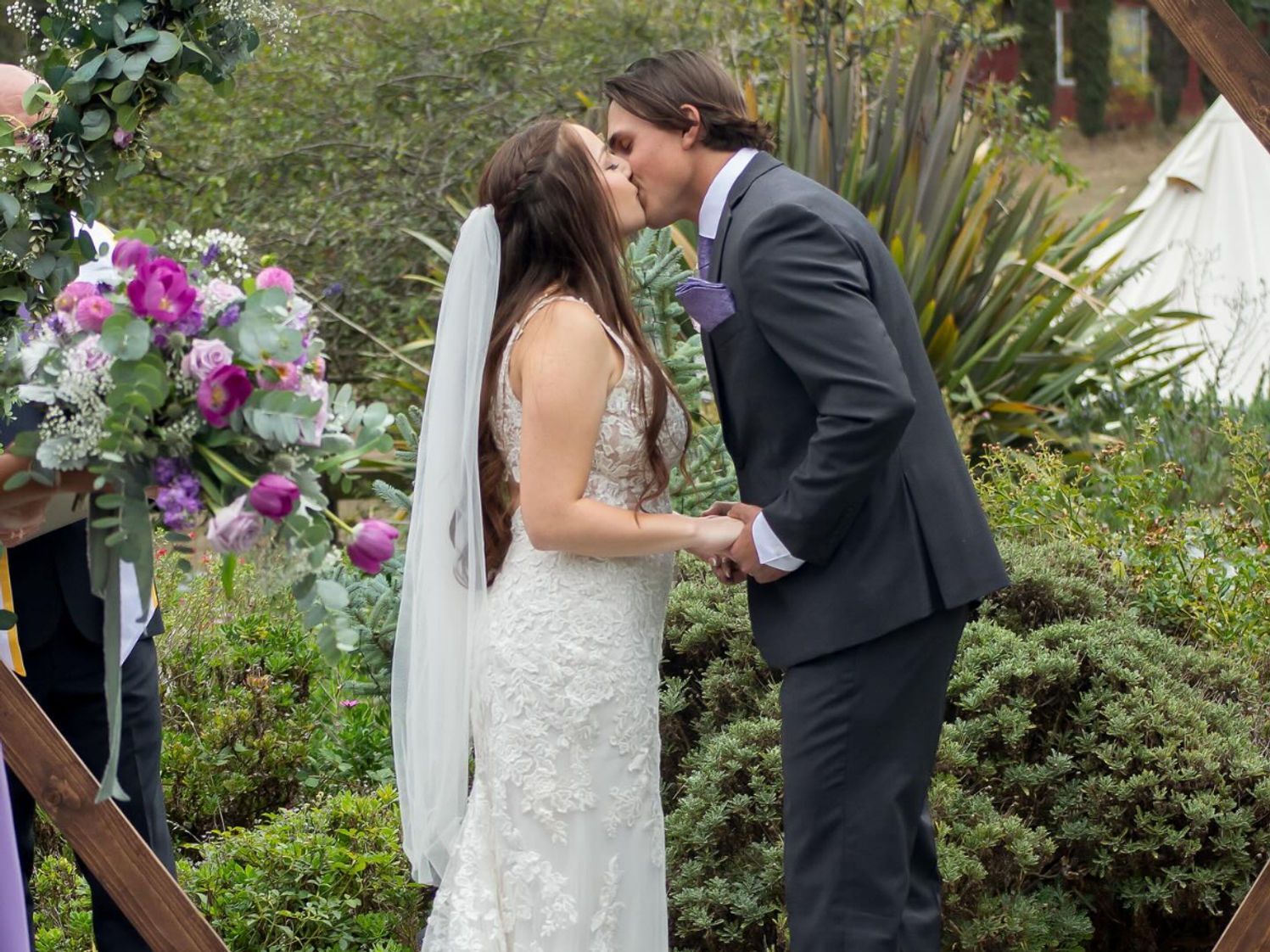 Bride and groom kiss at outdoor wedding ceremony; floral arch, holding hands.