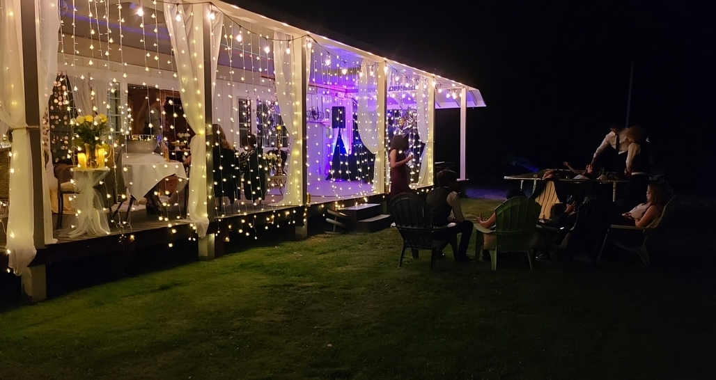 Nighttime porch decorated with lights. People sit in chairs on grass.