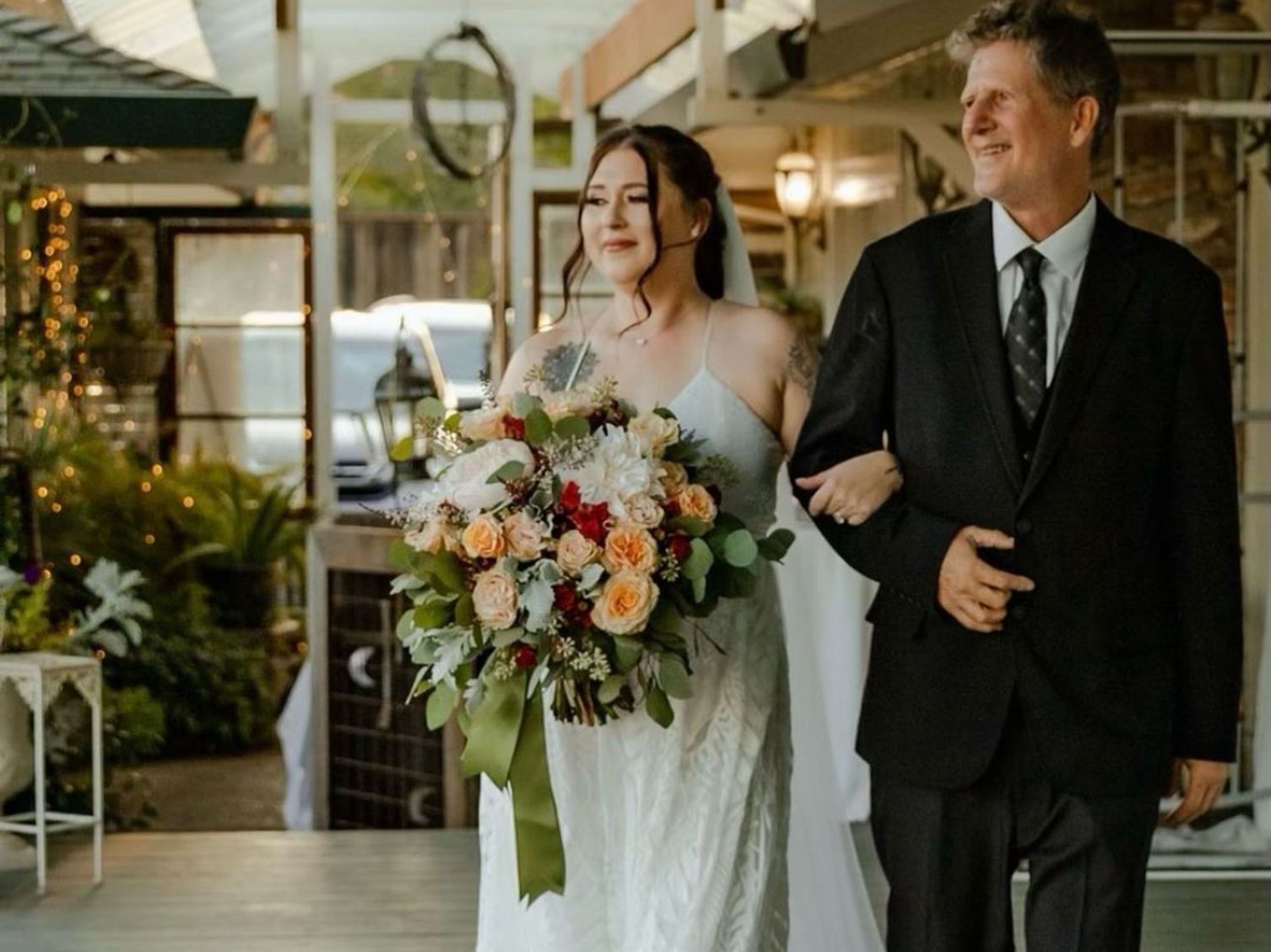 Bride walking down aisle with father; wedding ceremony. Bride wears white dress, carrying a bouquet. Father in black suit.