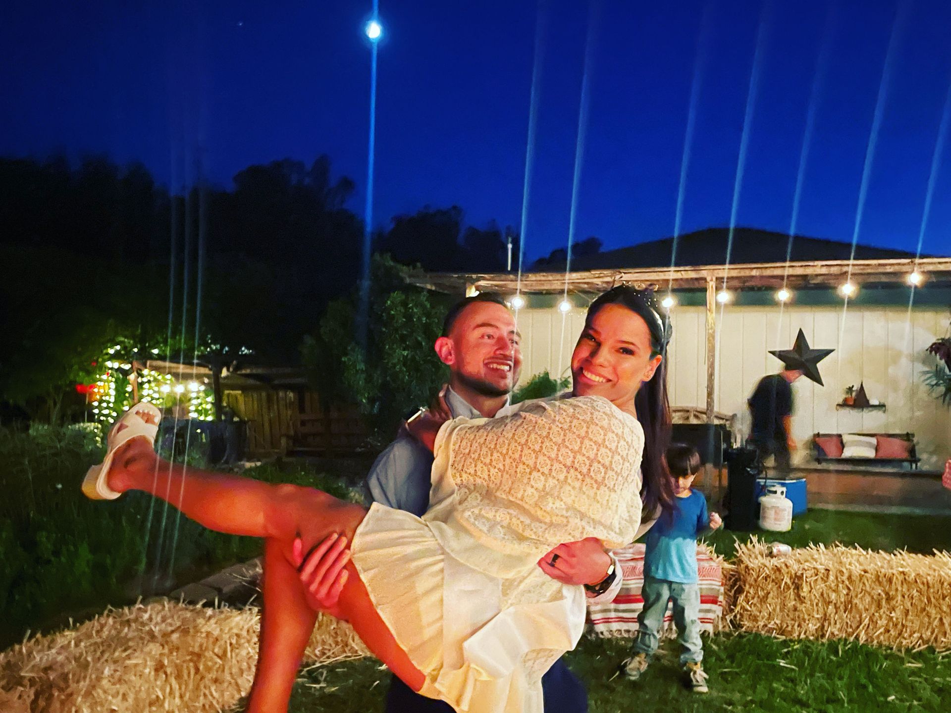Man holding woman, posing outside at night. Both smiling, backyard scene with lights and other people.