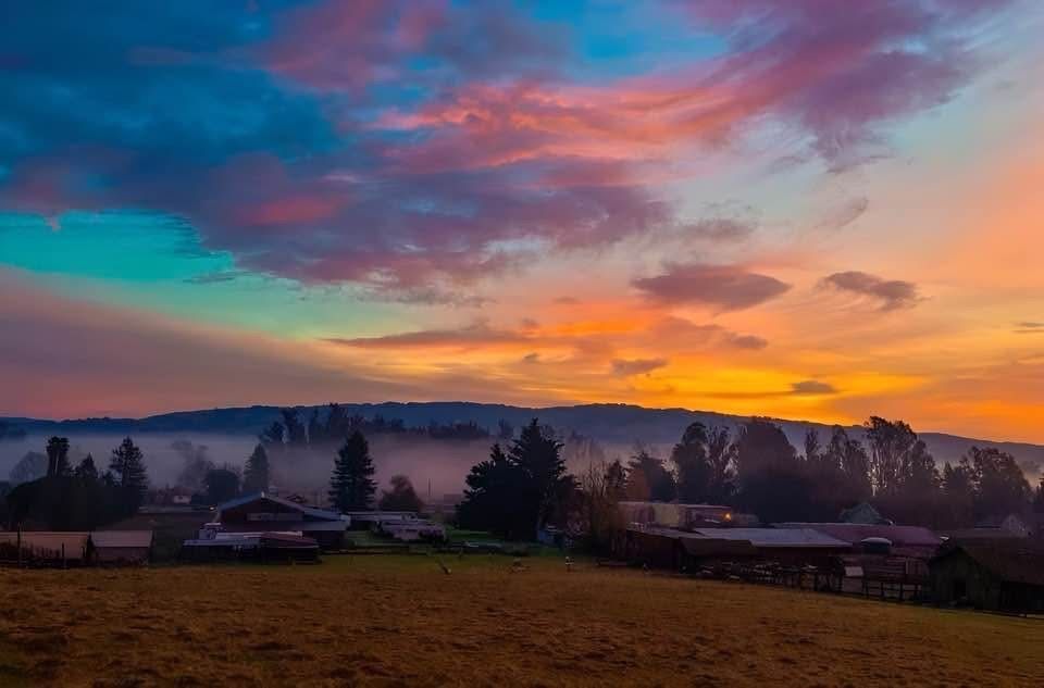 Sunset over a rural landscape; orange, blue, and pink sky above a foggy valley with trees and buildings.