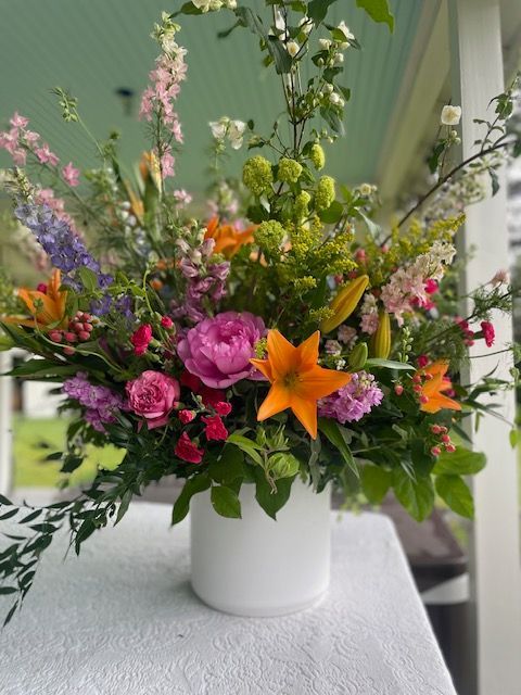 Colorful floral arrangement in a white vase on a white table outdoors; includes lilies and peonies.