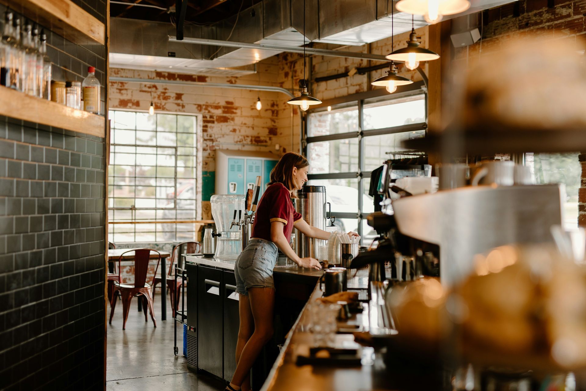 Woman in shorts behind a coffee shop counter, preparing a drink. Exposed brick, natural light.
