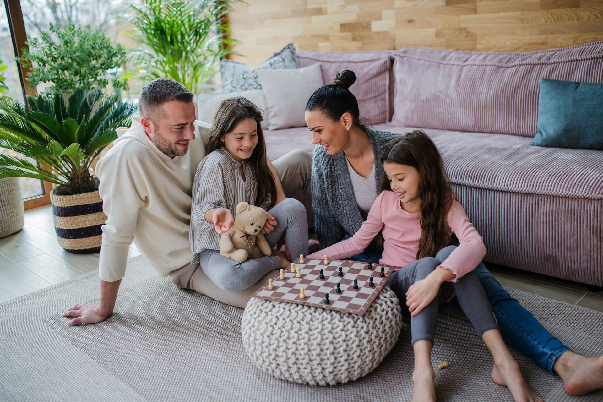 Family playing chess on a rug in a living room, smiling.