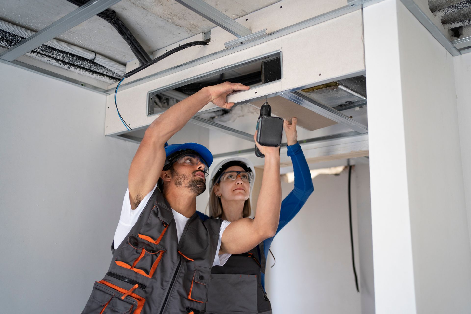 Two construction workers installing a ceiling panel with a drill. They are inside a building with exposed metal framing.