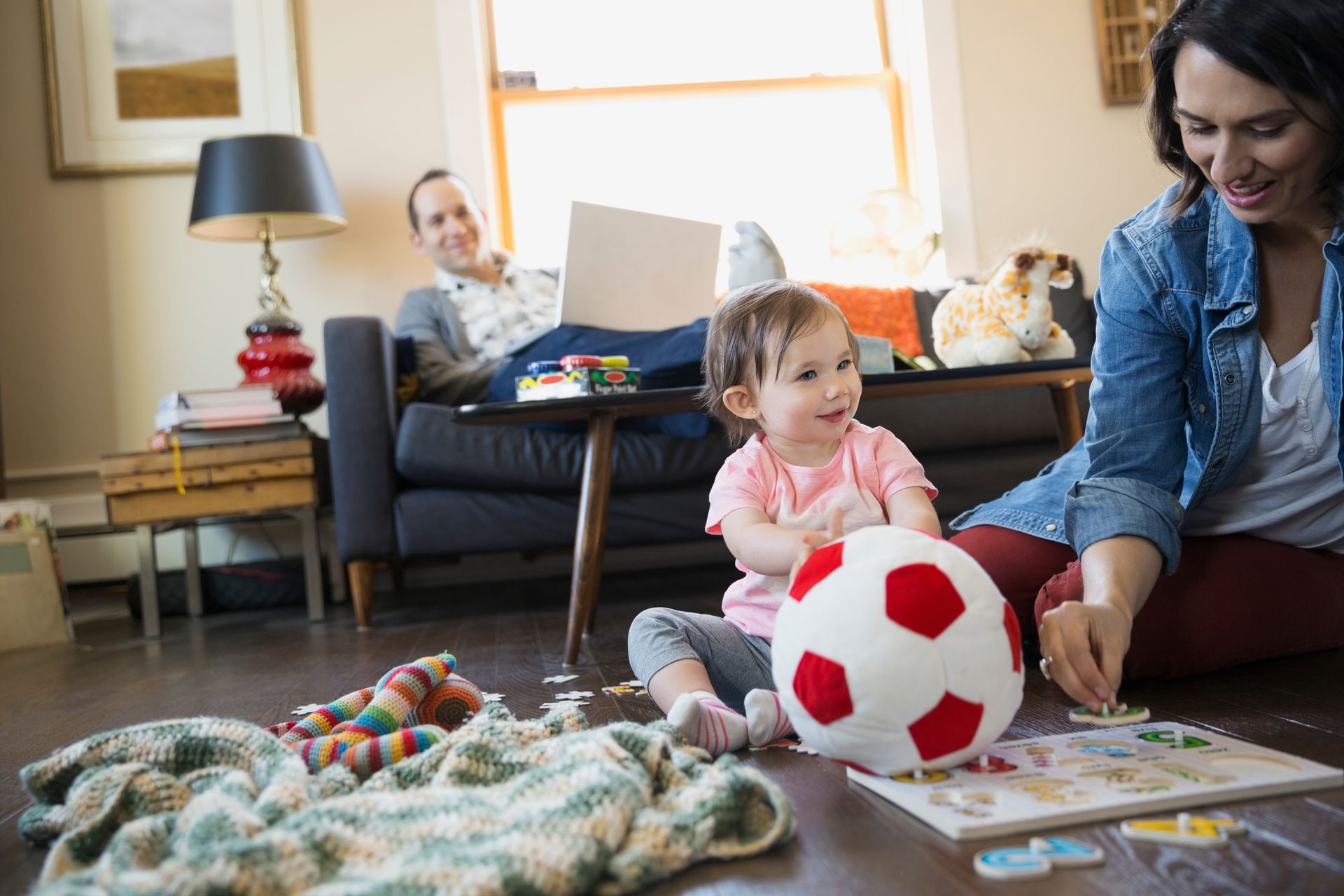 Woman and child playing with a ball and puzzle on the floor; man works on a laptop in the background.