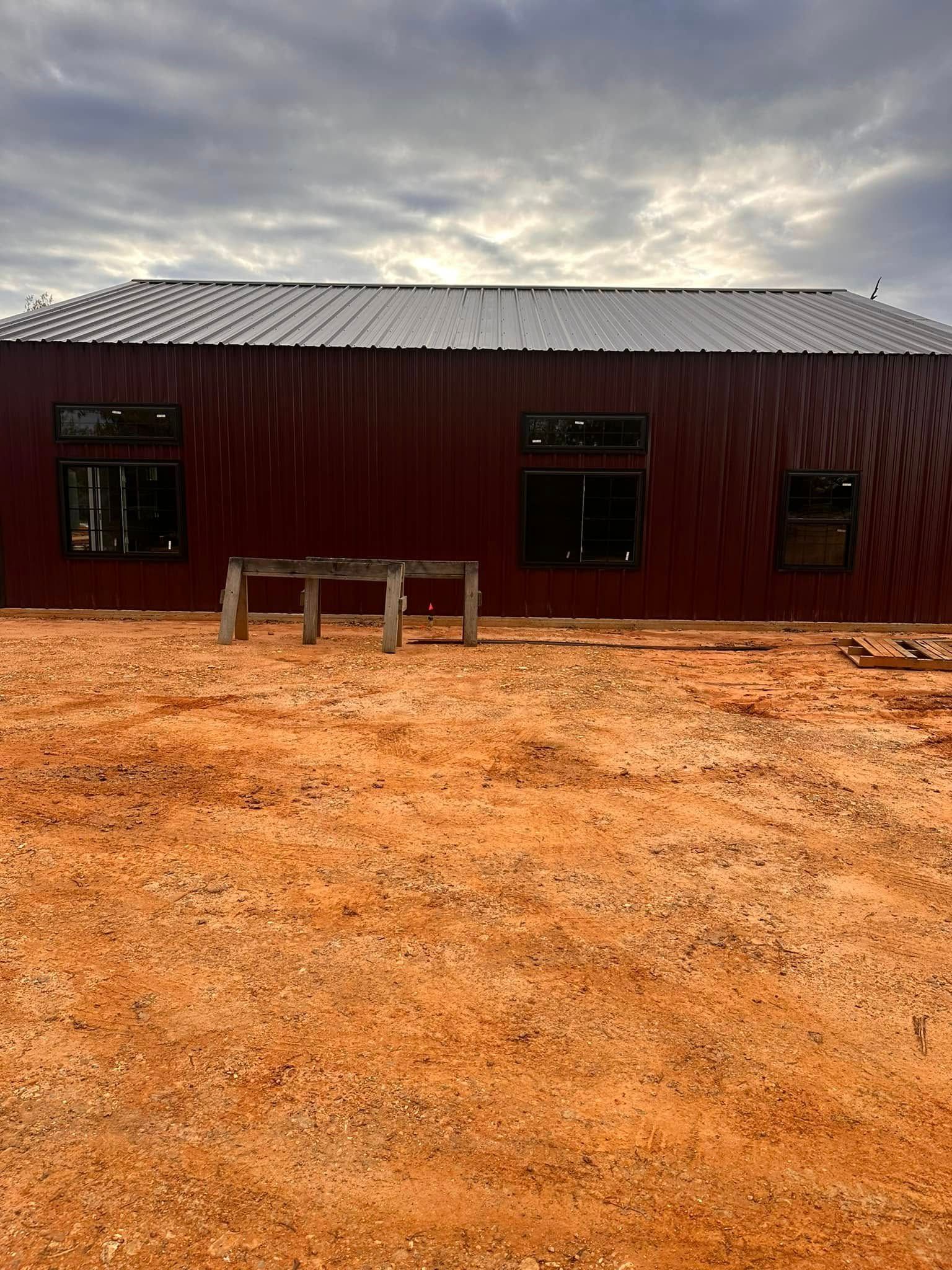 Red wooden building with a metal roof on a dirt lot under a cloudy sky.