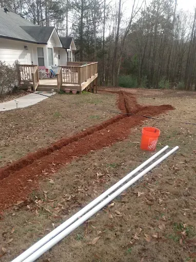 Trenches dug in yard, white pipes, orange bucket, wooden deck, house in background.