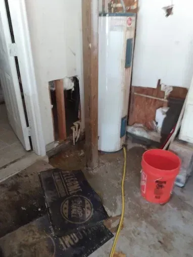 Water damage in room; hole in wall with pipes; water heater, bucket, flooded floor.