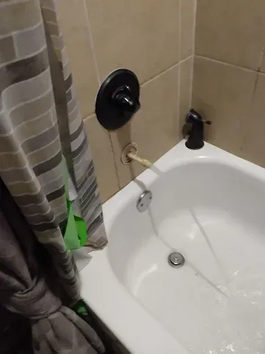 Bathtub with shower fixtures, exposed pipe. Dark faucet, shower curtain, beige tile wall.