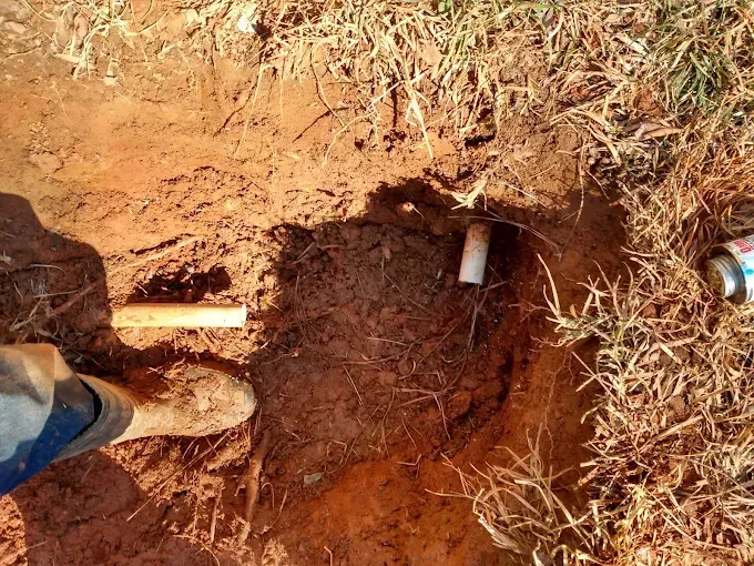 A hole in the ground with a PVC pipe and a foot visible; red dirt and grass surround.