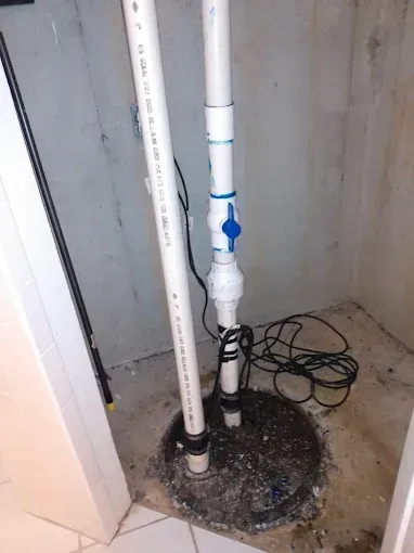 A sump pump system in a basement, with two white pipes, a valve, and a black electrical cord.
