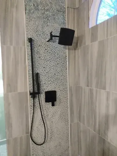 Black shower fixtures against a tile wall. Pebble-like accent tile strip.
