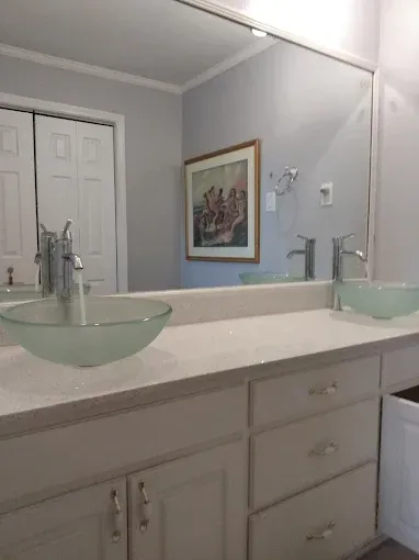 Bathroom with two glass bowl sinks, silver faucets, and a large mirror reflecting a painting and doors.