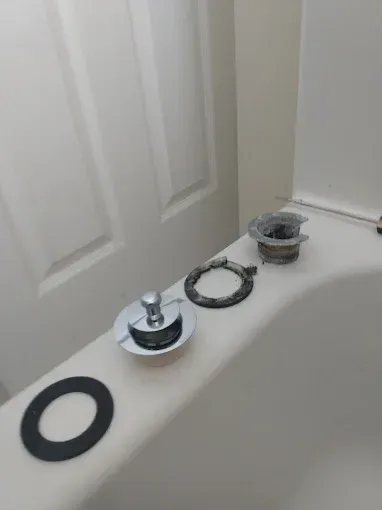 Bathroom tub drain parts disassembled on the tub rim. Includes a drain stopper and gaskets.
