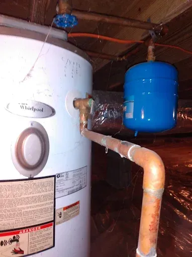 A Whirlpool water heater with a blue expansion tank connected by copper pipes in a crawl space.