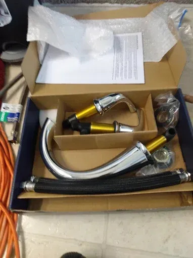 Box of chrome faucet parts, including handles, spout, and hoses, on a beige surface.