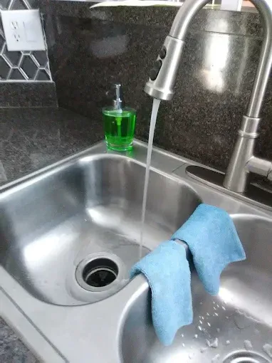 Stainless steel kitchen sink with running water, blue cleaning cloth, and soap dispenser.