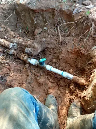Man in boots in a dirt trench, plumbing pipe with blue valve exposed.