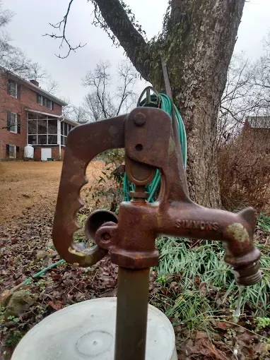 Rusty outdoor water pump with green hose near a tree and a brick house.