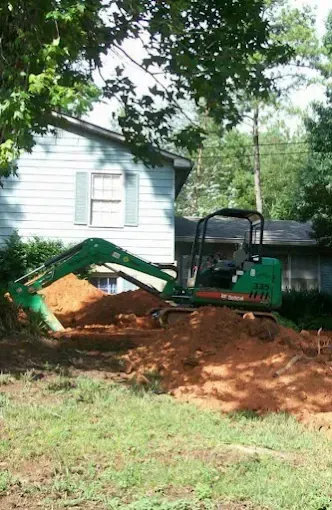 Green excavator digging dirt in front of a light blue house with shuttered windows. 