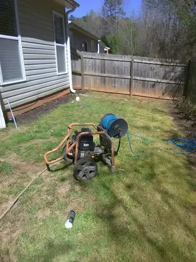 A pressure washer sits on grass near a house, hose extending toward a wooden fence.