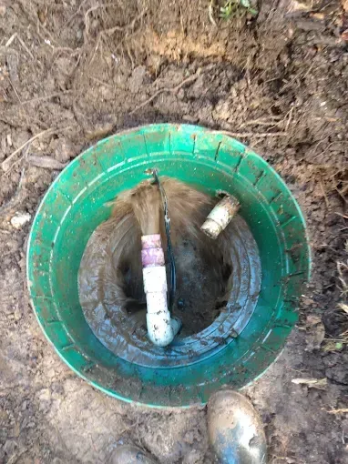 Open view of a green septic tank access point with exposed PVC pipes and muddy surroundings.