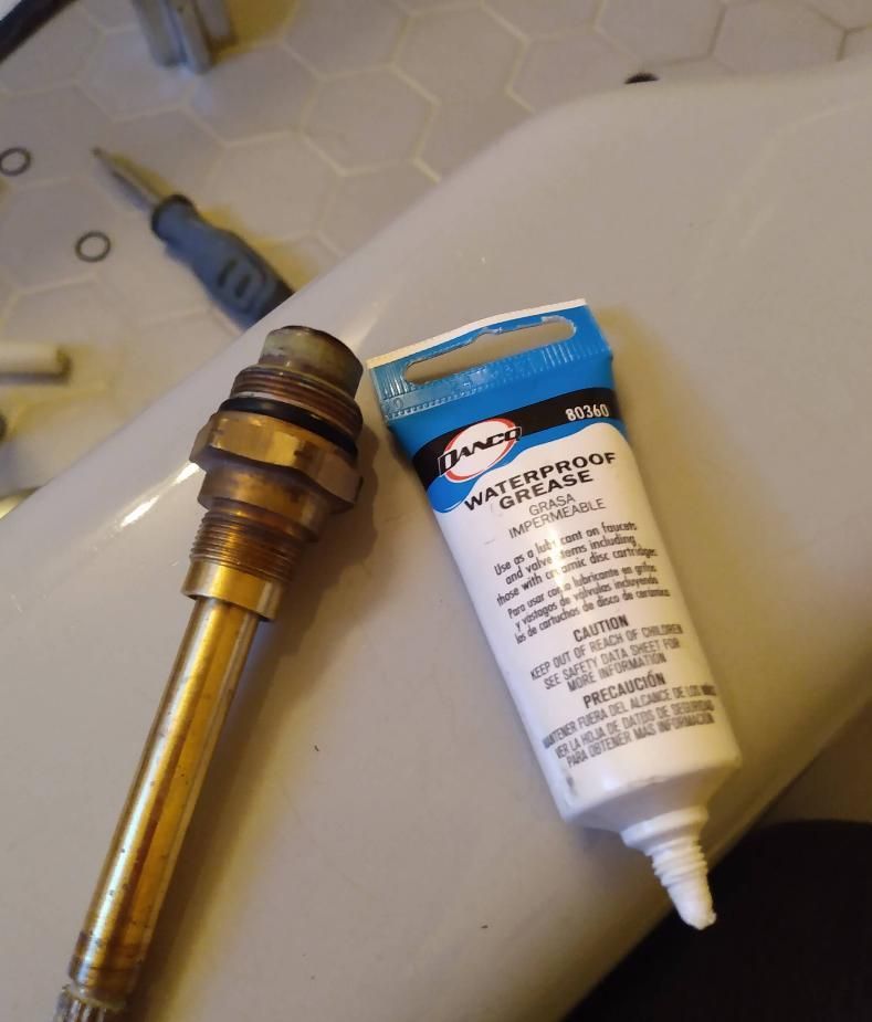 Brass faucet stem and tube of waterproof grease on a white bathtub rim.