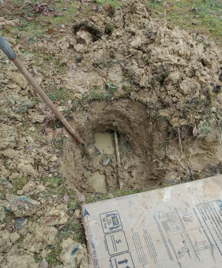 Hole dug in muddy ground, revealing pipe. A shovel leans against the side.