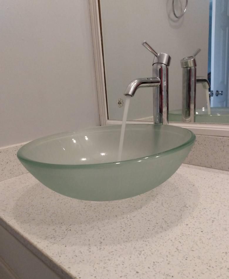 A glass bowl sink with a chrome faucet running water, sitting on a white speckled countertop.
