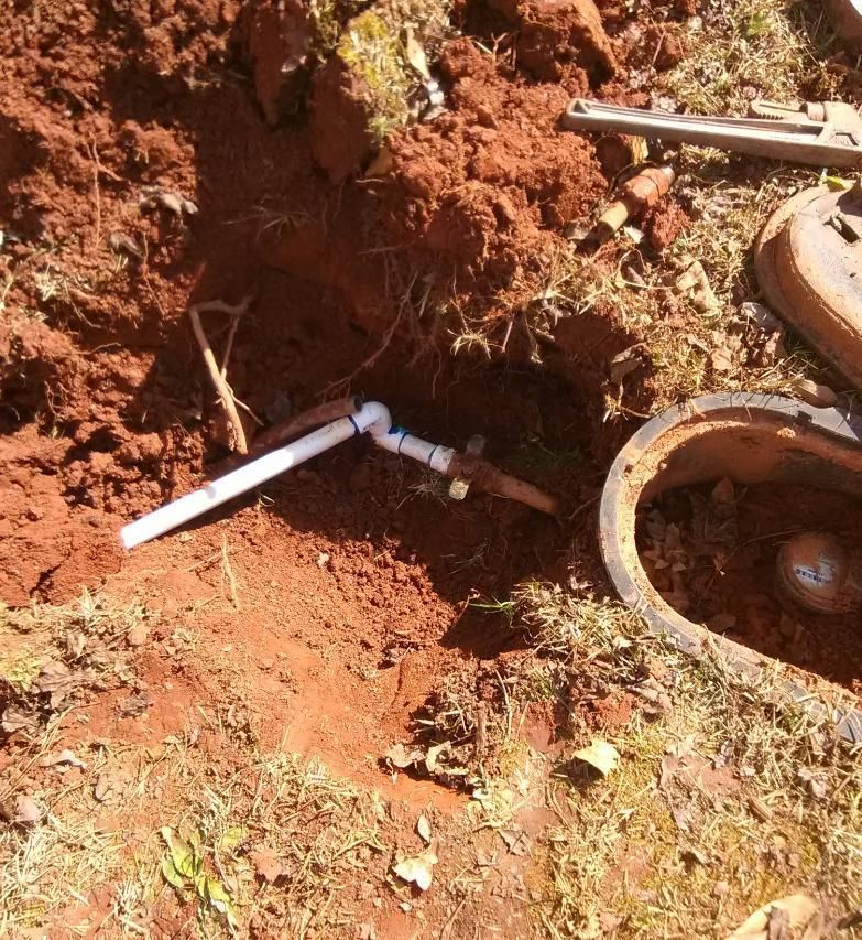 Red soil surrounds a buried PVC pipe and a water valve, near an open metal access lid.