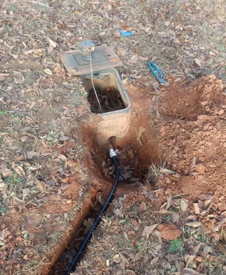 Open utility box in a dirt hole; black pipe runs from it along a trench.