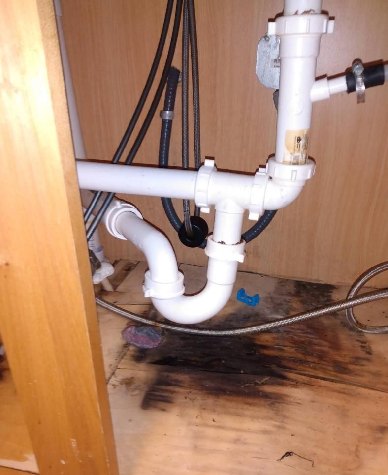 White sink pipes under a cabinet, with a visible water leak and mold on the wooden floor.
