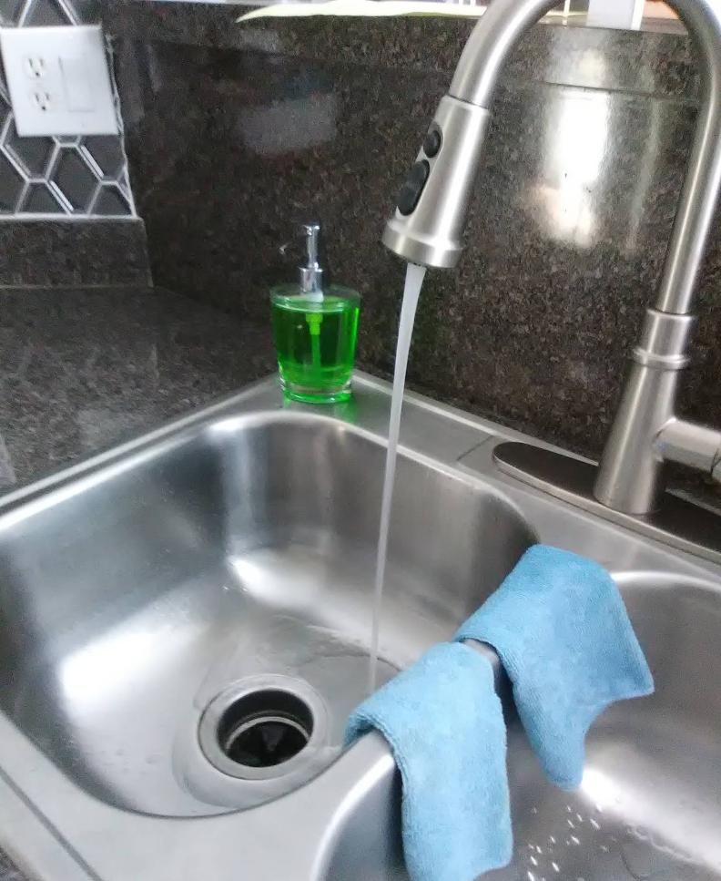 Stainless steel kitchen sink with running water, soap dispenser, and blue cloths.