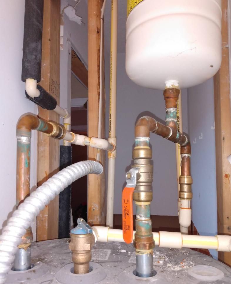 Plumbing pipes, including copper and PVC, attached to a water heater in a framed wall setting.