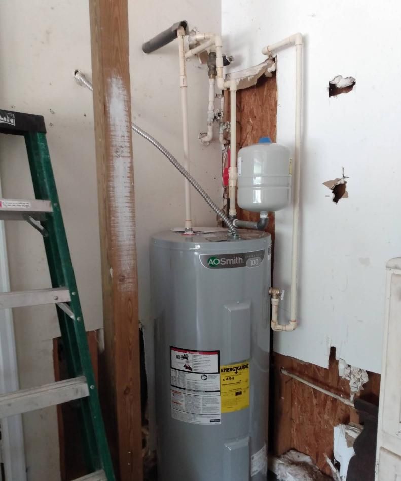A water heater in a damaged wall corner with exposed plumbing, beside a leaning ladder.