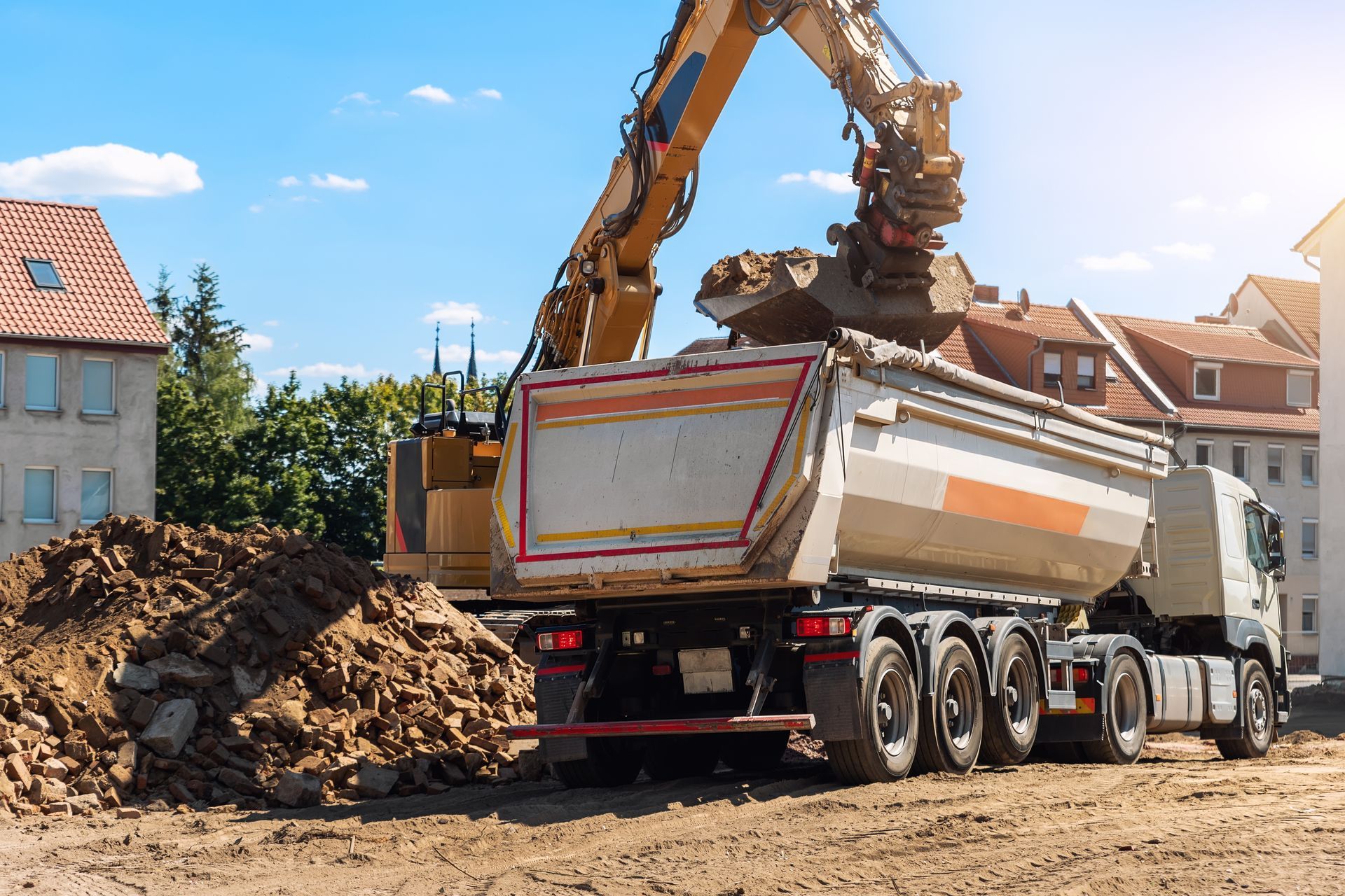 Excavator loading dirt into a dump truck at a construction site, in front of buildings on a sunny day.