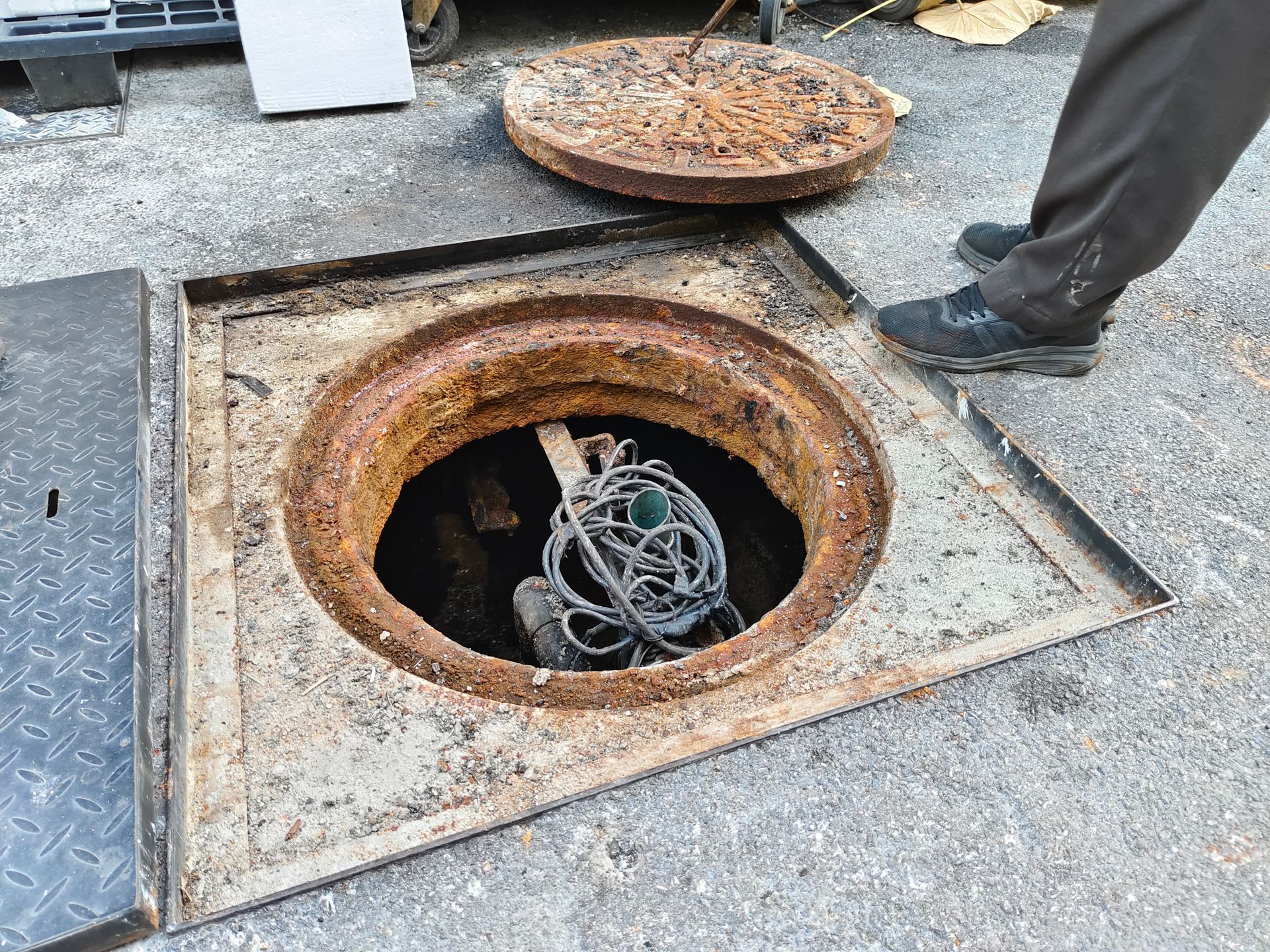 Open manhole with wires inside; person standing nearby. Rusty metal, concrete ground.