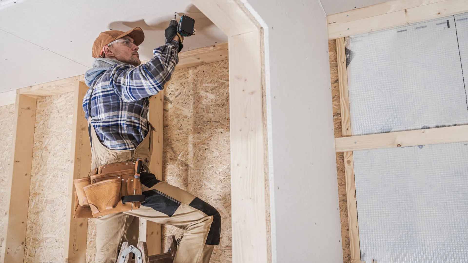 Un ouvrier du bâtiment installe des plaques de plâtre dans une pièce, debout sur une échelle.