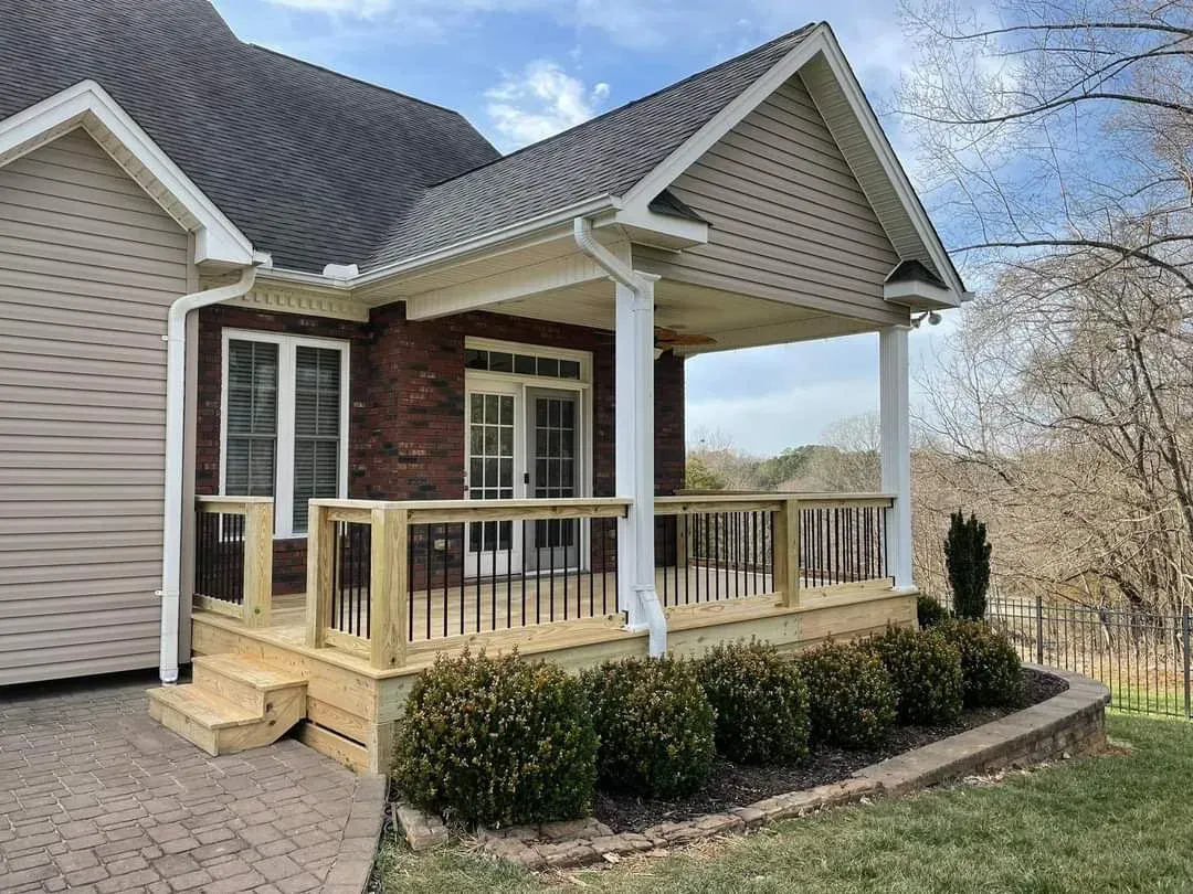 Brick house with a covered porch, wooden deck, and a small garden bed with green bushes. The house has a brown roof and is surrounded by a paved driveway and grass.