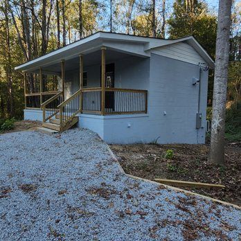 Small light blue house with a porch, wooden railing, and gravel driveway in a wooded area.